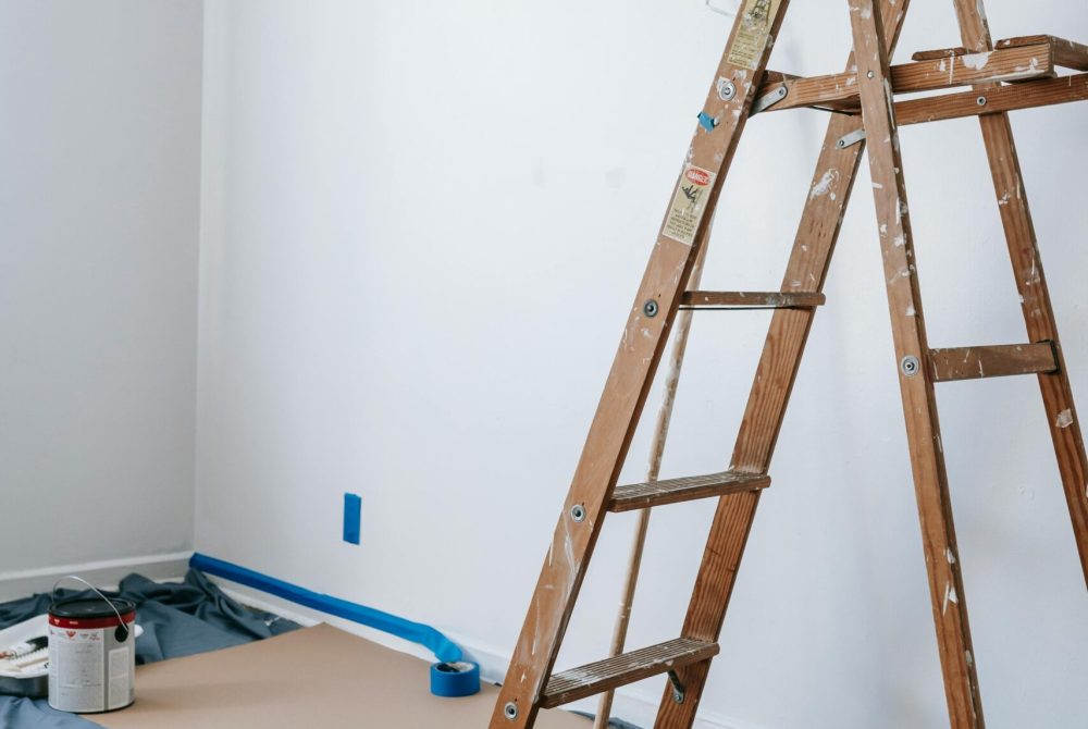 A home renovation scene featuring a wooden ladder, painting tools, and white walls in a room.